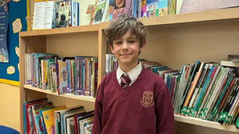 Harry Bavington stood in his school library with books on the shelves behind him. He is wearing his maroon school uniform jumper with a white shirt and maroon tie.
