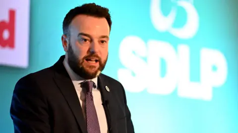 Colum Eastwood speaking in front of a backdrop that has SDLP branding. He is wearing a suit and tie