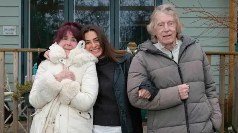Solène Declas Three people are standing in front of a summer house wrapped up in thick coats smiling to the camera. Solene Declas is standing in the middle, with her arm around her grandmother Marion Riley on the left and her other arm is linked with her grandfathers, Joe Riley. Marion has dark red hair that is tied up. Solène’s hair is long and dark brown. Joe has fair hair which is long enough to touch his collar.