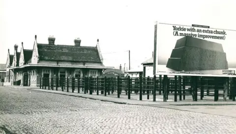 Arthur Johnson and Sons A black and white photo showing livestock pens in a cobbled area in front of counting house buildings. A large advertising board for Yorkie chocolate is on the right of the image.