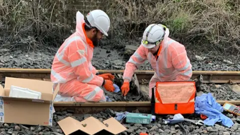 Network Rail Two men wearing white safety helmets and orange overalls sitting on a train track carrying out repairs. There are boxes and tools around them.
