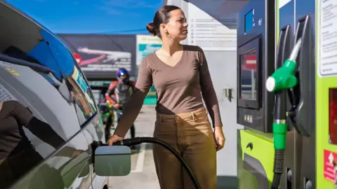 A woman wearing a brown long sleeved top and beige trousers filling up her car at a petrol station on a sunny day