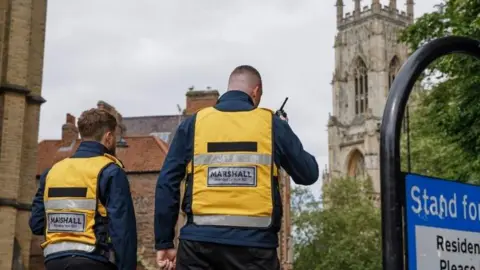 The York BID The back of two men wearing yellow hi-vis jackets over navy coats and black trousers. One is talking into a walkie-talkie. They are walking around the city of York, near the minster. 