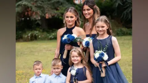 Family photo Ruby is wearing a navy bridesmaid dress and holding blue and white flowers. She is surrounded by her three sisters, who are wearing matching dresses, and next to her twin brothers, who are wearing light blue shirts.