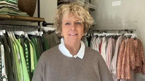 Leanne Rinne/BBC The owner of Nice Boutique in Rye, Lesley Farrow, stood in front of two rails of clothing stock. Lesley has dyed blonde hair and is wearing a grey jumper and white undershirt. 