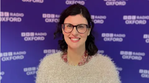 A photo of Layla Moran MP in front of purple branding for Radio Oxford. She is wearing dark rimmed glasses and a fluffy beige jumper. 