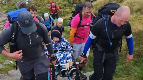 Eight people going up Ben Nevis, in climbing gear with equipment, carrying Ted in his wheelchair, he is wrapped in blue, white and white clothes.