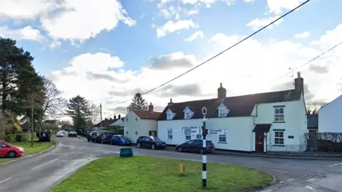 Google View of the village green in Bassingham with a black and white fingerpost direction sign in the foreground and a white cottage in the background.