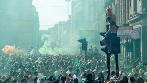 A large gathering of fans on the Trongate in Glasgow. Green and orange smoke fills the air, mostly green, and people sit up on top of traffic lights. People wear Celtic strips and wave Celtic flags 