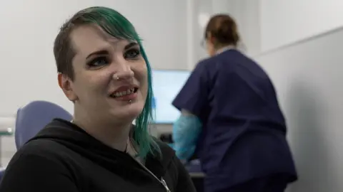 A woman sits in a chair as a health worker in scrubs stands behind her. 