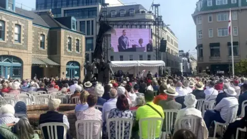 BBC People are sat down at a previous event. They are sitting down on white chairs and looking at a large screen. There is buildings pictured around the side with a white tent at the front.