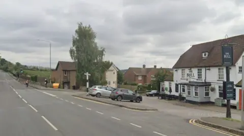 A Google street view image of a road. To the right there is a white pub called The Bull set back from the road.