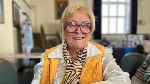 Clare Worden/ BBC Hillary Letch has a blonde bob and red glasses. She is wearing a white shirt and yellow gilet.