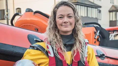 RNLI/Simon Culliford Tamara Brookes is standing in front of a RNLI rigid inflatable boat and is smiling at the camera. She has long, curly blonde hair and is wearing her RNLI uniform and buoyancy aid.