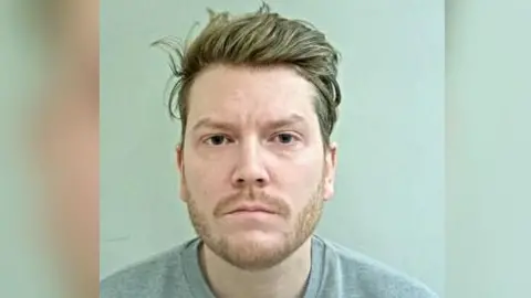 A police mugshot shows Jacob Gill looking at the camera with no facial expression. He has short light brown hair and a light brown moustache and beard. He is wearing a grey top.