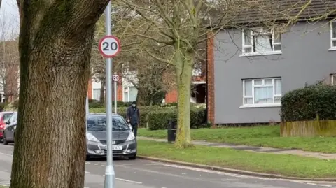 A road in Stevenage, showing 20pmh signs, a man walking, parked cars, houses, flats and a grass verge by a road. 