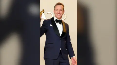 A man wearing a black tuxedo is holding up an Oscars trophy