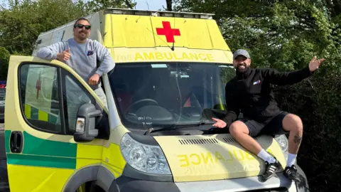Rhys Edwards hangs out the driver's side of an ambulance and smiles at the camera with his thumb up, while Jake Trask sits on the bonnet of the ambulance and smiles with his arms out.