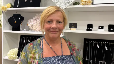 Heidi in a blue and white patterned dress and a patterned green and pink cardigan standing in front of jewellery. She has short blonde hair and smiles at the camera. 