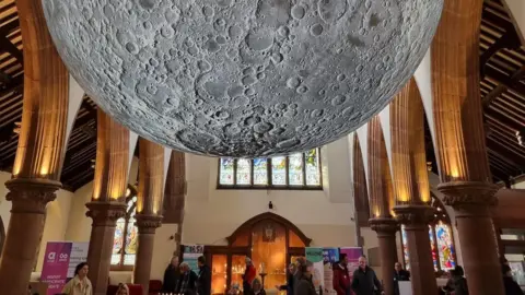 A close-up of the bottom half of the suspended moon, which has visible craters, with the arches of the cathedral on either side.