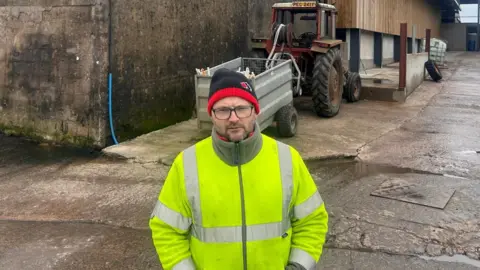 Ian Bowness is wearing a red and black woolly hat and a high-vis jacket. He has black glasses and a beard. He is standing on a farm, in front of a tractor.