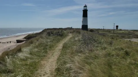 Harry Appleyard A lighthouse on a grassy cliff. A sandy beach and the sea can be seen to the left. The sky is blue and features light, white clouds. 