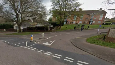 Road junction on right with bungalow on corner and low-rise block of brick flats off the road and a bus stop.