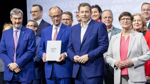 Getty Images Friedrich Merz in a blue suit and holding a document stands with his team of ministers - including three men in blue suits in the front and a woman in a white jacket