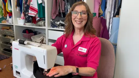 A woman with long brown hair and glasses is sitting at a sewing machine and smiling at the camera. She is wearing a pink collared shirt with the Dressability logo on it