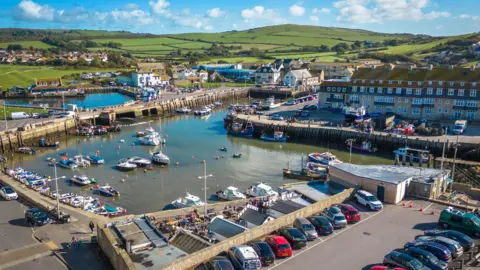 West Bay's small, square harbour has moored pleasure craft and open sides revealing fields and hills in the background.