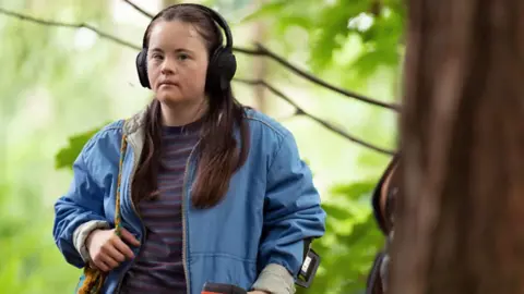 BBC/House Productions/Vishal Sharma A woman wearing headphones, a light blue jacket and a stripy top underneath. Her right hand is resting on the strap of her handbag