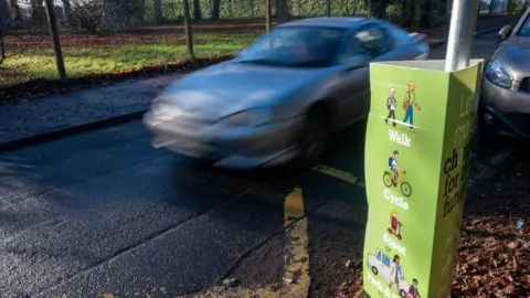 Green placard outside a schoolgate advising on methods of travel including cycling, walking and scooting. The sign is close to a road with a car driving past.