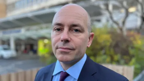 Steve Hubbard/BBC Roland Sinker, who is standing in front of a hospital. He is wearing a navy suit jacket, blue shirt and maroon tie.