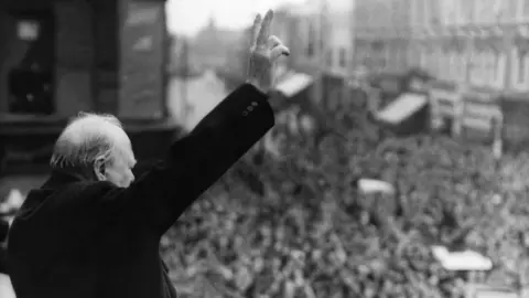 Getty Images A black and white photo of Winston Churchill making his famous victory sign to a crowd of people.