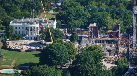 An aerial image of the White House, a large white building with tall classic columns. To the right of the building there's a large construction site, with scaffolding and cranes building a new structure. 