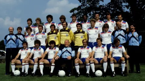 England's 1982 World Cup squad stood in three layers. They are all wearing England football shirts in different colours: white, blue and yellow. There are three footballs on the ground. 
