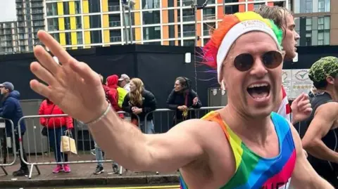 Family handout A man running a race is wearing a rainbow coloured outfit and hat, along with sunglasses. He is waving to the camera. 