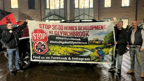 Three men stand beside a banner which two of them are holding up outside Forest of Dean District Council's offices on a wet night. The banner has a painting of a kingfisher sitting on a branch near a small stream in a field. There are some houses in the background of the banner, which reads "Stop 3500 homes development at Glynchbrook".