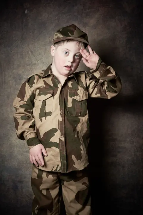 Debbie Todd Maximus, a young boy with short blond hair salutes the camera as he wears camouflaged trousers, jacket and hat