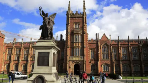 The exterior of Queen's University Belfast, a large gothic redbrick building with a statue in the font of an angel. There are some people 