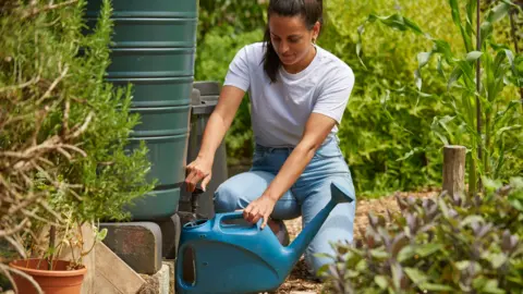 Thames Water A woman in a white T-shirt and blue jeans filling up a watering can from a water butt. She is crouched down with greenery all around her.