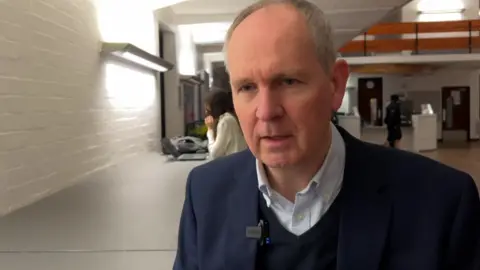 Professor Rowan Sutton looking at the camera is wearing a white shirt, navy v-neck sweater and a blue suit jacket. He has grey hair and blue eyes.