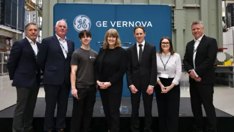 GE Vernova Seven people stand in a large factory, in front of a stage covered in a black material, which is in front of a blue sign which says "GE Vernova".
