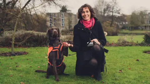 Dr Elena Ratschen Dr Elena Ratschen, a woman wearing a black coat and a pink scarf, is crouching down on grass, holding the paw of a brown dog that is sat beside her