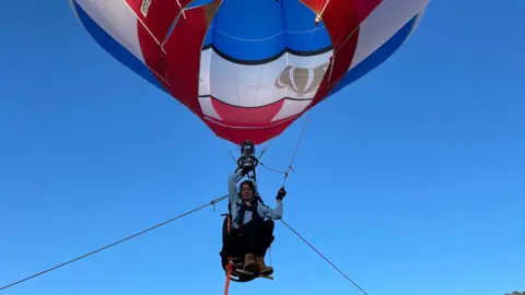 Jo Bailey is sat in a single person hot air balloon in the sky with ropes tied on either side. 