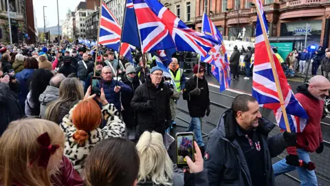 Pacemaker A procession waving UK and Israel flags walk down a city street. A crowd of onlookers in winter attire can be seen recording the procession on their phones. 