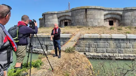 A man stood outside a grey Victorian sea fort. He is being filmed by a person using a camera on a tri-pod. Another man is stood behind the person filming.