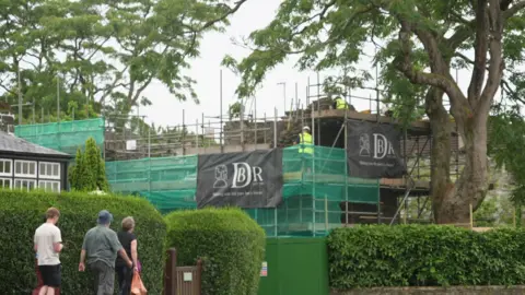 The medieval Betsy Grimbal's Tower is covered in scaffolding and green safety netting during conservation work, with specialist builders working on the structure above a public footpath.
