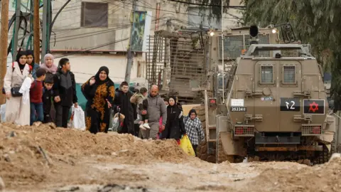 Reuters Palestinians walk next to Israeli military vehicles and an armoured bulldozer on a destroyed road from Jenin refugee camp, during an operation by Israeli security forces, in the occupied West Bank (22 January 2025)