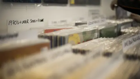 WeArePlaster A close-up of shelves filled with vinyl records. The vinyls are packed in closely together and all have plastic wrapping on them. They are split into sections, with one at the back reading '1970s and later, soul + funk'.